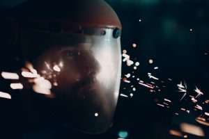 Some of the most common hazards in factories that can cause eye injuries include: ● Flying objects: this can include metal chips, wood chips, dust, and other small particles. Adult bearded man in transparent protective mask and grinder saw with flying metal particles sparks in darkness